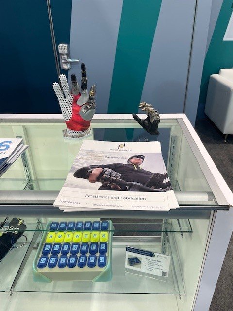 Two prosthetic hands sitting on a showcase display cabinet infront of flyers from Point Designs. 